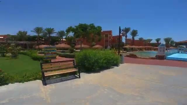 I giardini del Caribbean World Hurghada con il prato verde, una panchina in legno all'ombra e la piscina con gli ombrelloni di paglia sullo sfondo