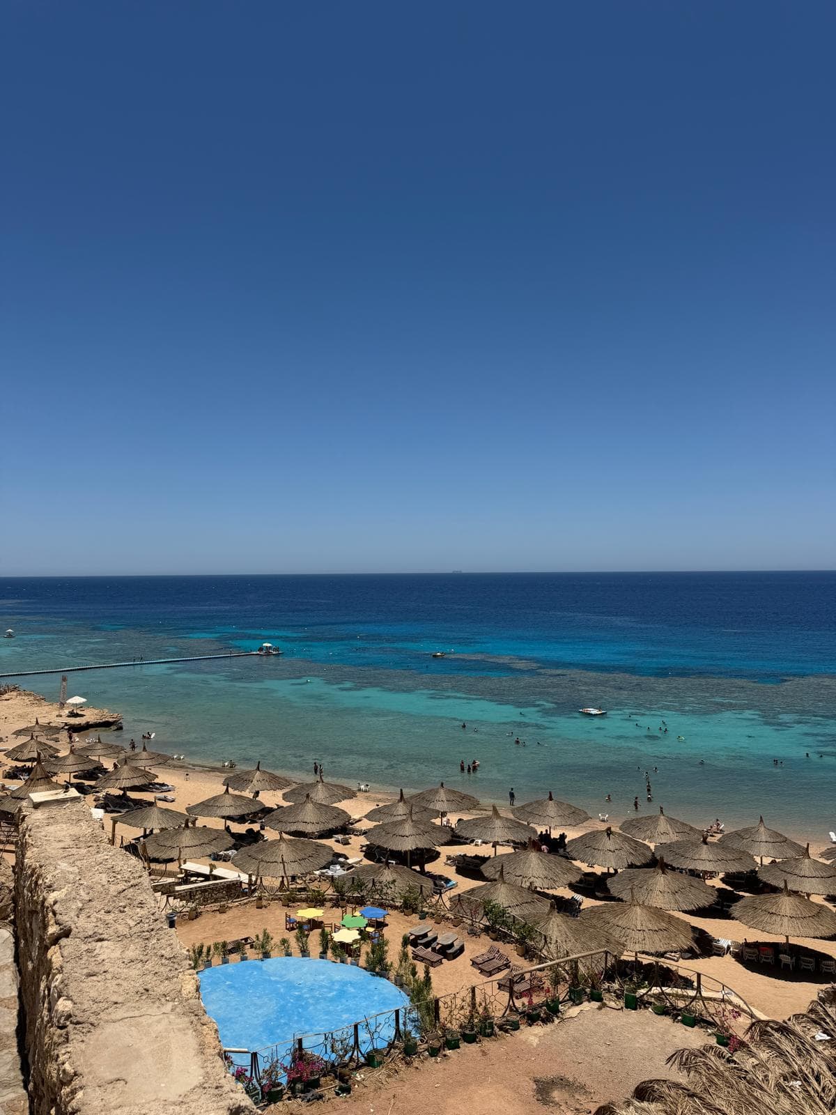 La baia di Ras Um Sid vista dall'alto: ombrelloni di paglia sulla sabbia, l'acqua turchese bassa e il disegno scuro della barriera che si legge sotto il pelo dell'acqua, con il pontile che la attraversa