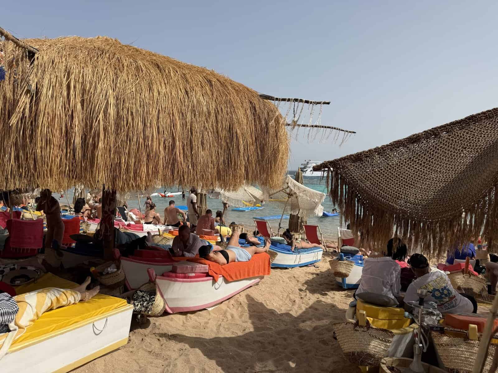 Un beach club di Sharm in una giornata piena: lettini e cuscini colorati sotto le tettoie di paglia, gente in acqua e una barca all'ancora