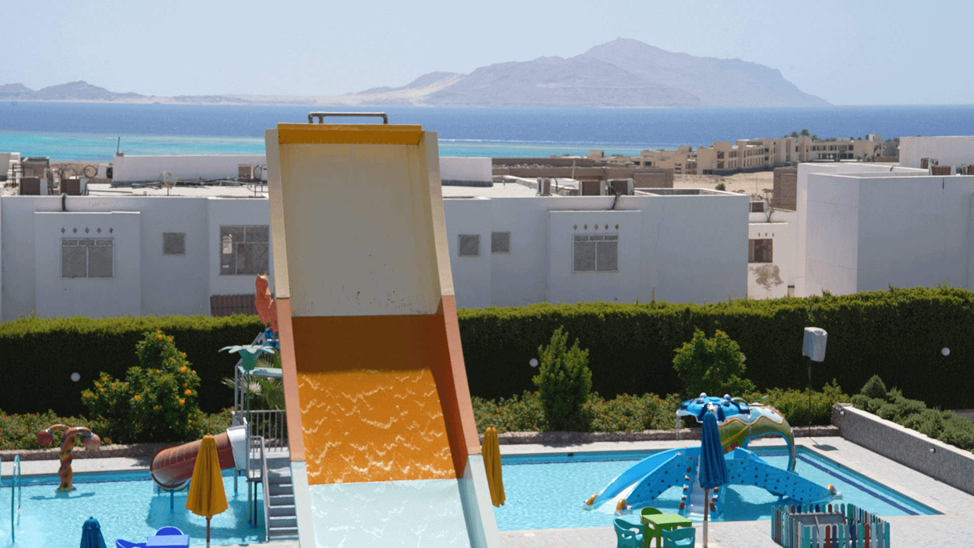 Parco acquatico con scivolo arancione, piscine blu e resort bianco con vista sul mare e isole in lontananza