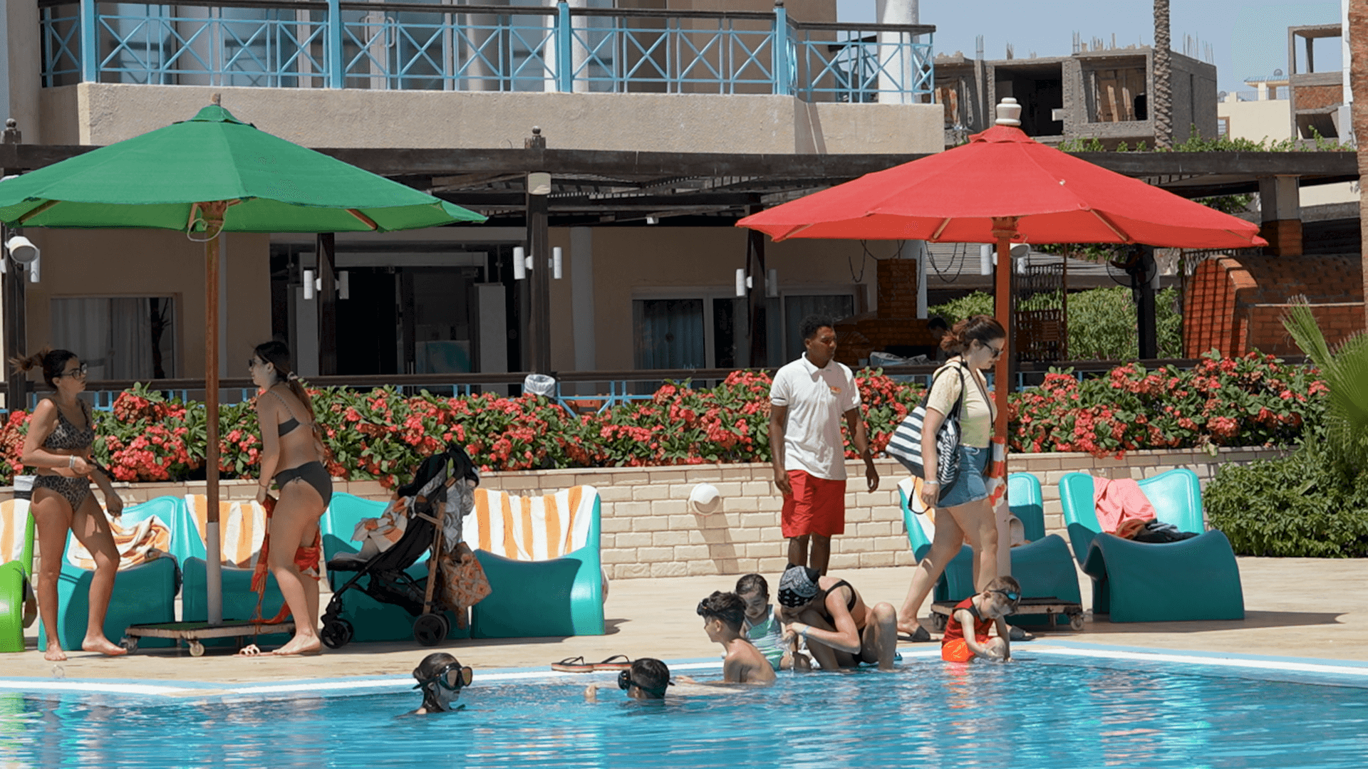 Persone in costume da bagno in una piscina con bambini che giocano in acqua, ombrelloni colorati verde e rosso, lettini blu, fiori rossi e edifici moderni sullo sfondo