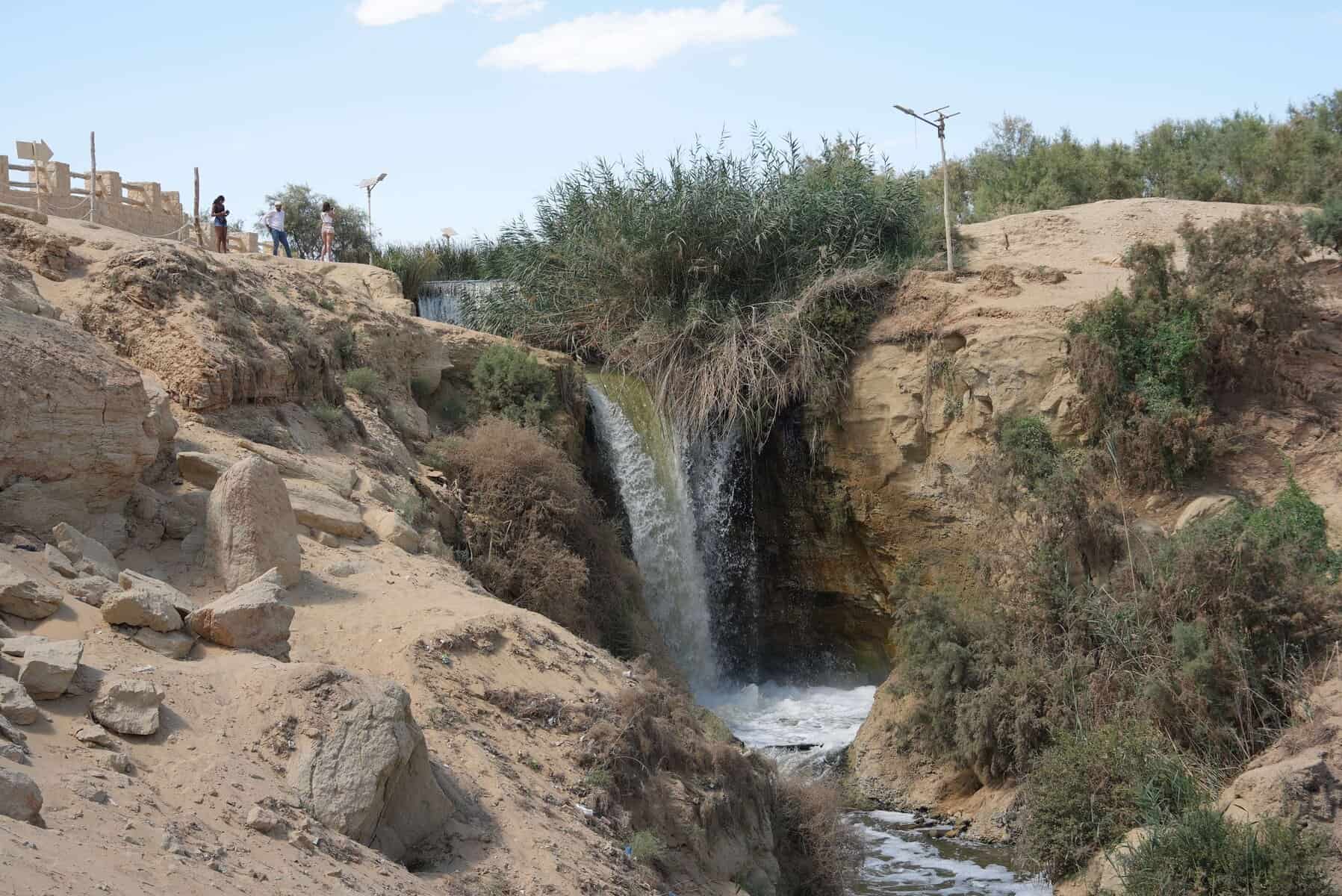 Cascata in una gola sabbiosa del deserto egiziano con vegetazione lungo le sponde e persone in visita sulla riva superiore