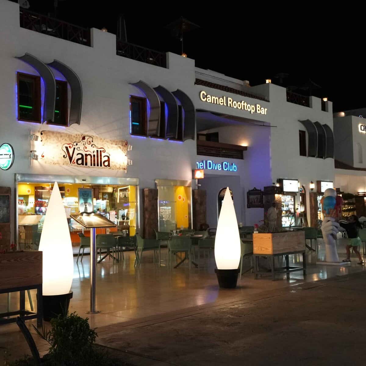 Piazza notturna con bar e locali illuminati: Vanilla, Camel Rooftop Bar e Camel Dive Club con lampade decorative bianche