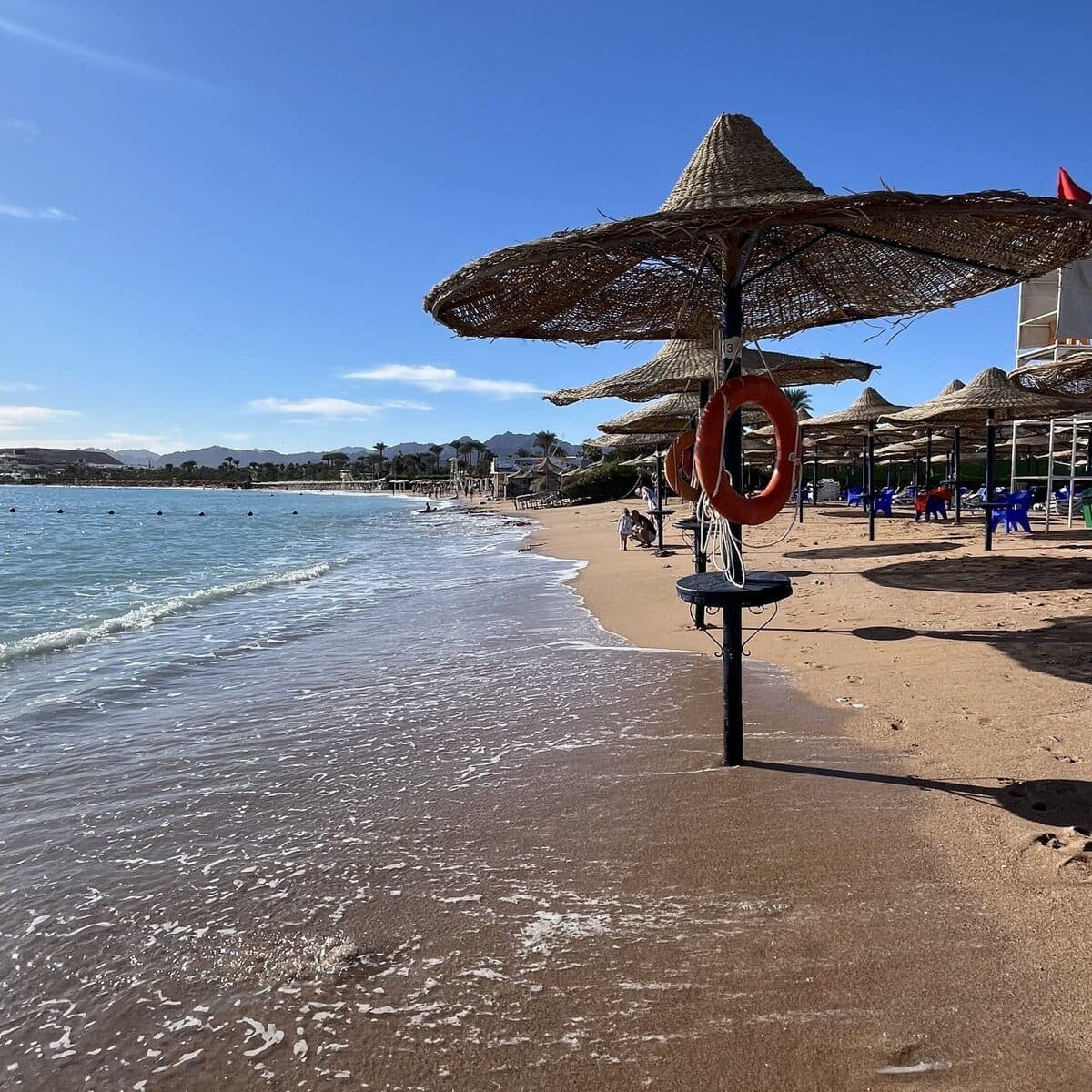 Spiaggia di Sharm in primavera: ombrelloni di paglia con i salvagente appesi, acqua calma e trasparente, le montagne del Sinai sullo sfondo