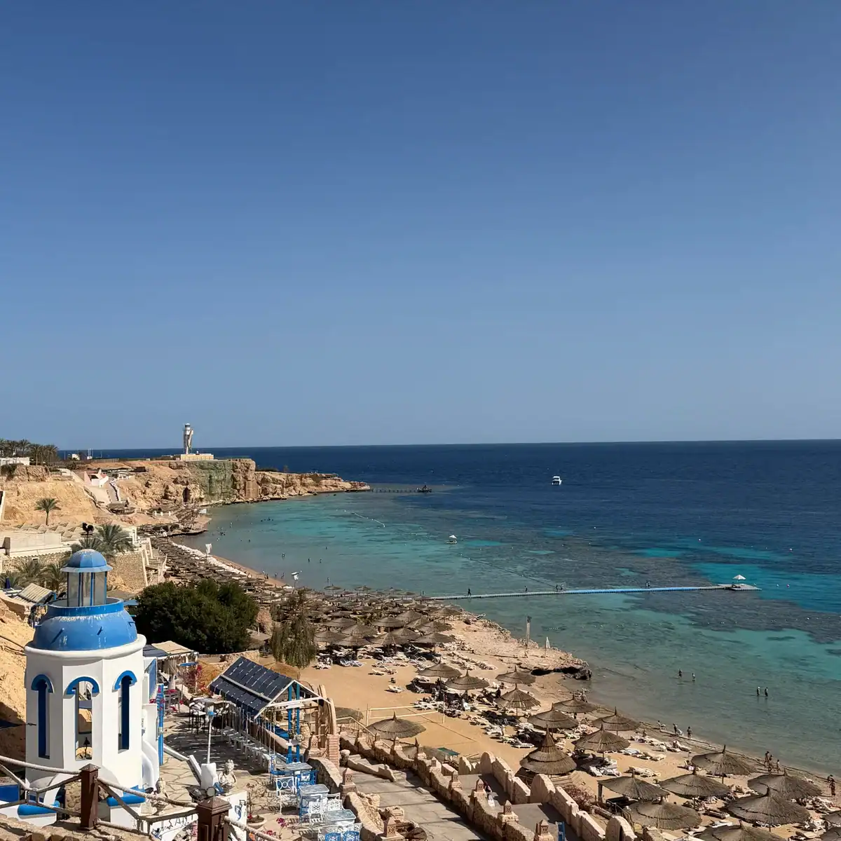 La baia di Ras Um Sid vista dall'alto in una giornata d'inverno: cielo limpido, mare piatto e turchese, il pontile che attraversa la barriera e gli ombrelloni di paglia sulla spiaggia