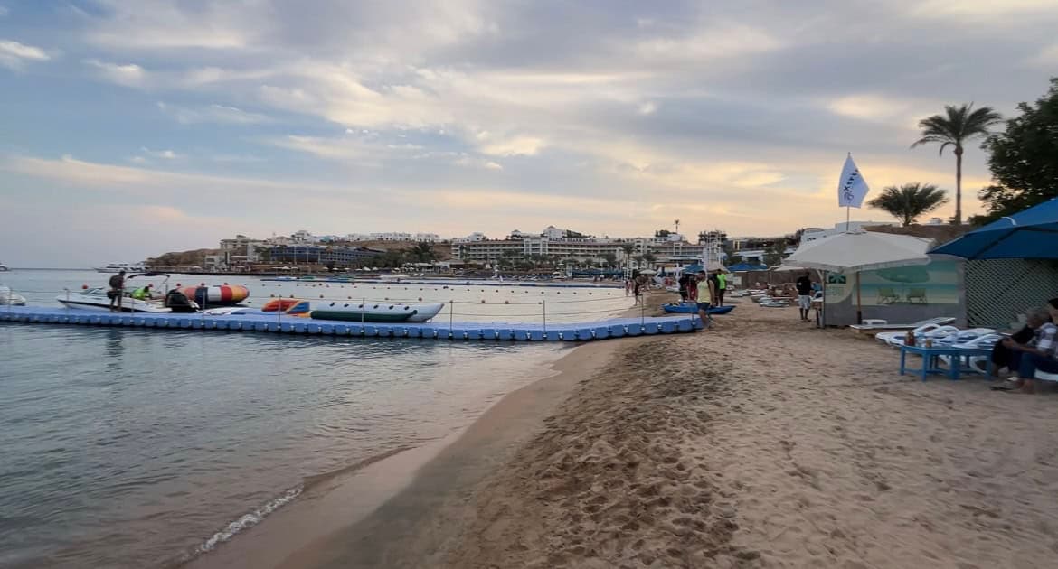 Spiaggia con zone di balneazione delimitate da cordoni blu, resort sullo sfondo, palme e ombrelloni sulla costa sabbiosa