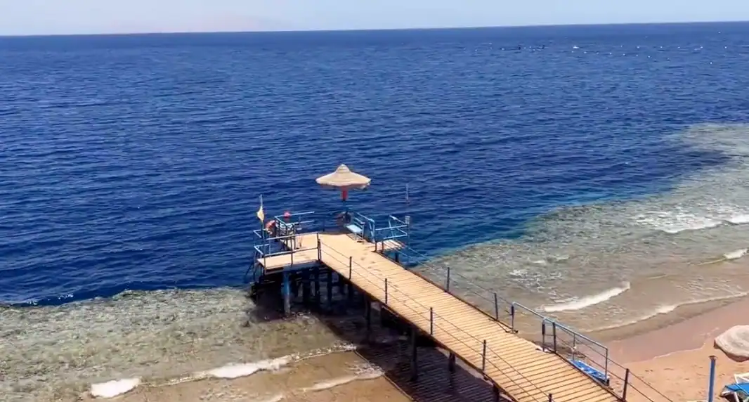 Pontile in legno con ombrellone beige che si estende in acque blu cristalline di una spiaggia egiziana, con sabbia dorata e onde leggere