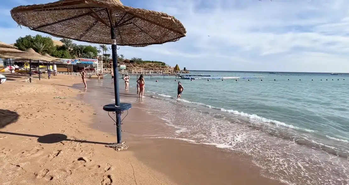 Spiaggia sabbiosa con ombrellone in primo piano, bagnanti in acqua cristallina e strutture balneari sullo sfondo