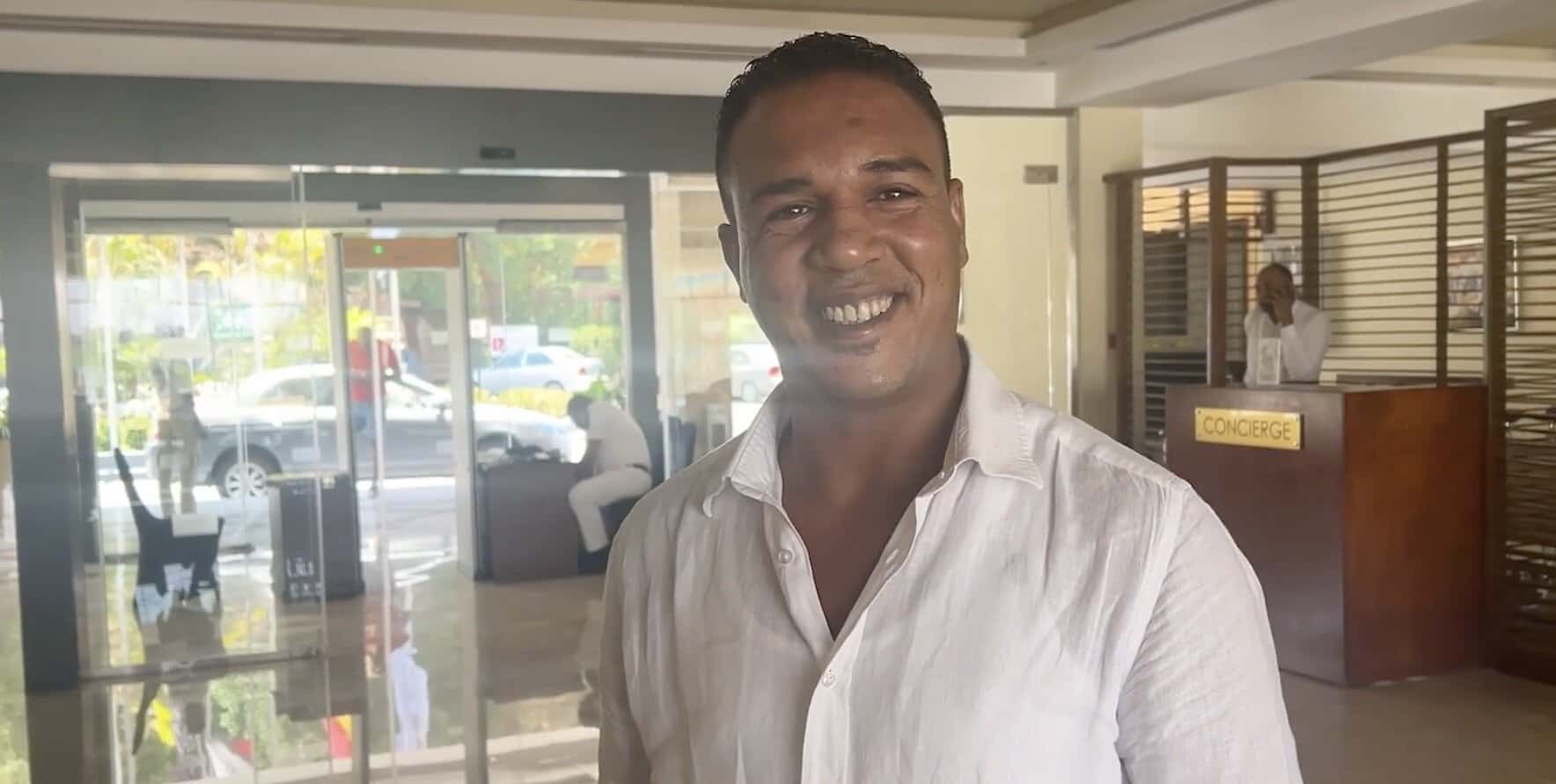 Uomo sorridente in camicia bianca in una hall di hotel con concierge, porte in vetro e arredamento moderno sullo sfondo