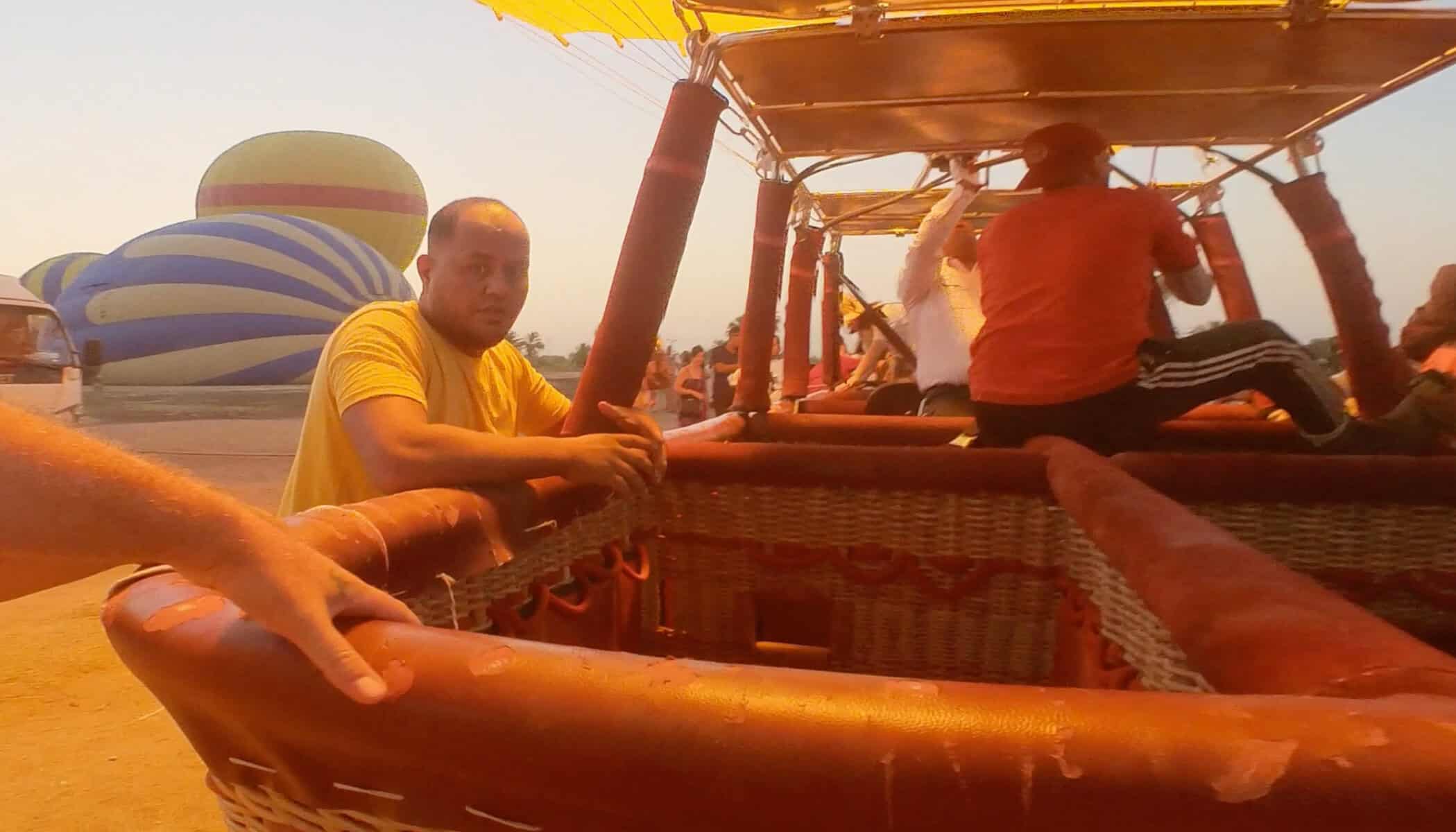 Dentro un cesto di mongolfiera al tramonto con persone che controllano le corde, sullo sfondo un'altra mongolfiera blu e gialla
