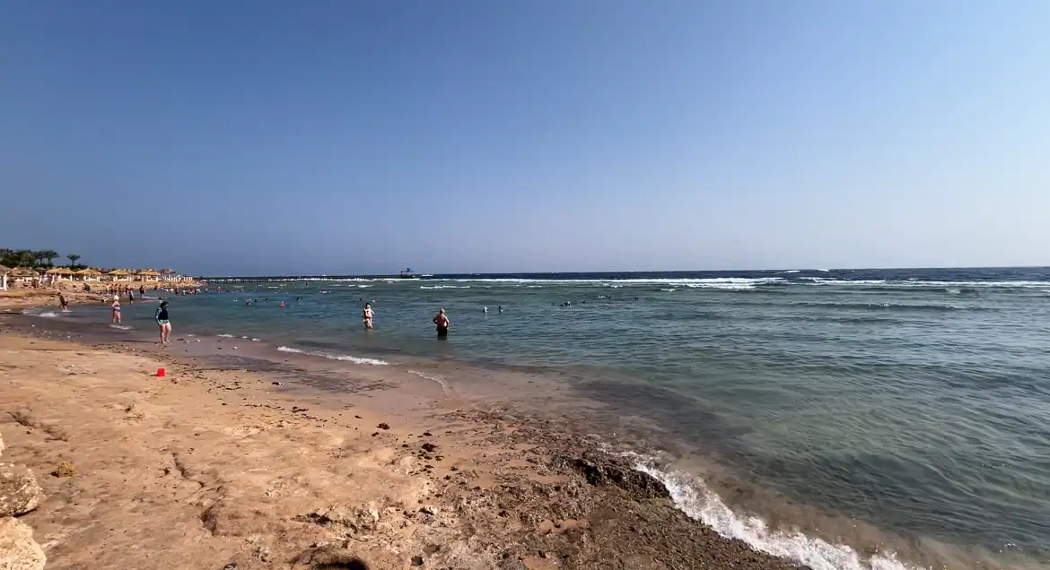 Spiaggia di sabbia dorata con bagnanti in acqua turchese, resort e ombrelloni sullo sfondo sotto cielo sereno
