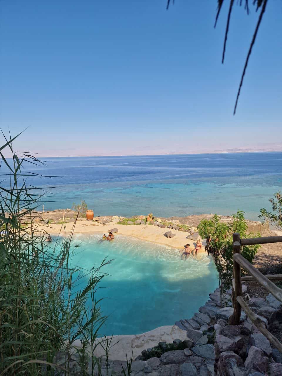 Sorgente d'acqua termale turchese con bagnanti sulla spiaggia del Mar Rosso, circondata da rocce e vegetazione desertica