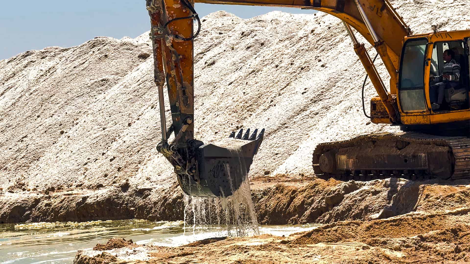 Escavatore giallo che scava con la benna in un cantiere di estrazione, mucchi di sabbia bianca sullo sfondo e operaio nella cabina
